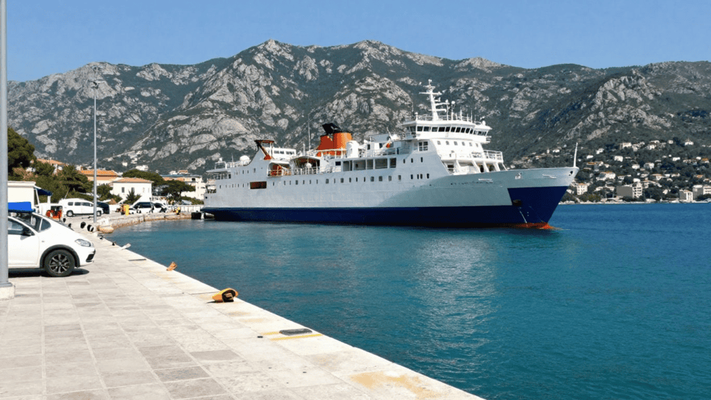 A large white cruise ship docked at a marina with turquoise water and mountains in the background.