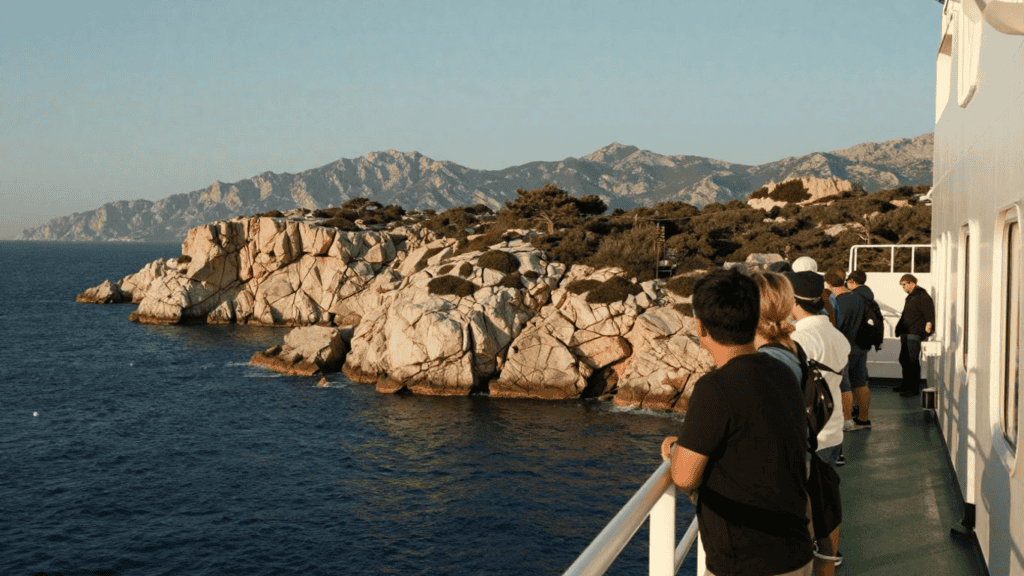 Passengers on a ferry deck looking out at rocky coastline and distant mountains during sunset/sunlight