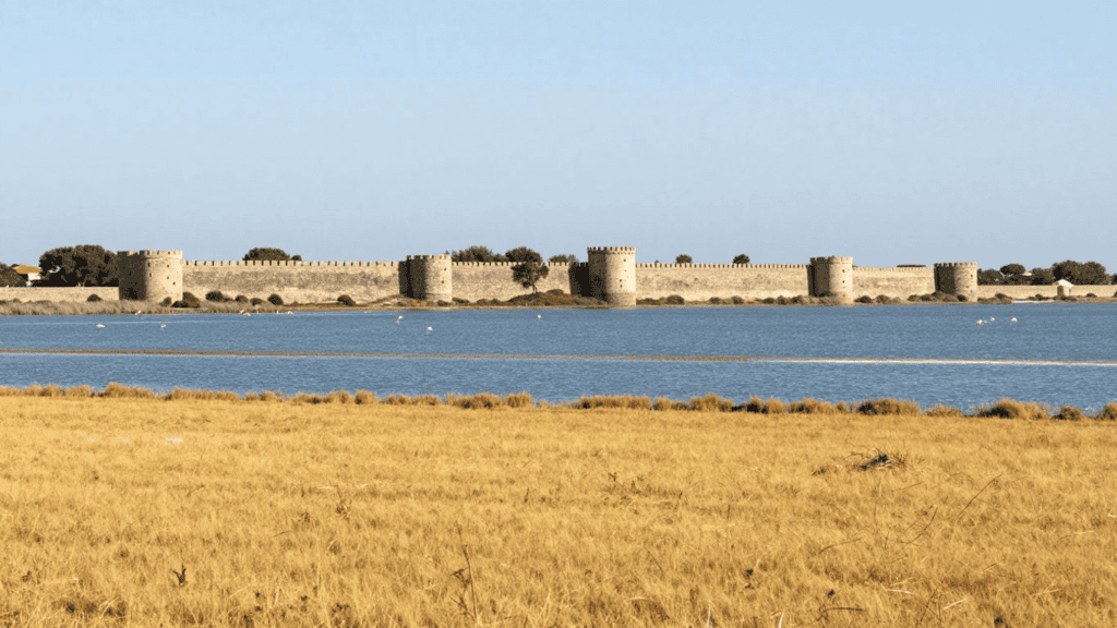 Stone fortress wall with round towers along a calm blue waterway, dry grassy foreground under a clear sky.
