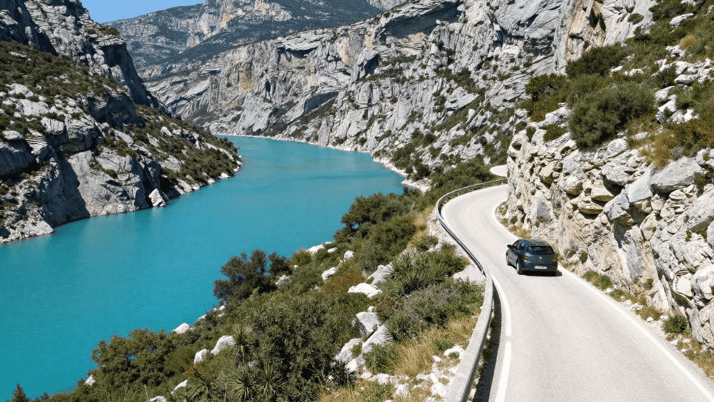 Winding coastal road along a turquoise mountain lake with steep rocky cliffs on both sides.