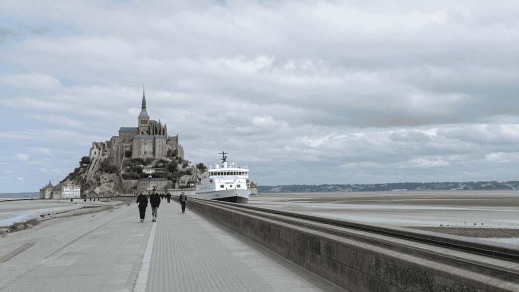 Mont Saint-Michel abbey on a rocky island with a long causeway, a white passenger boat docked nearby, and people walking along the walkway.