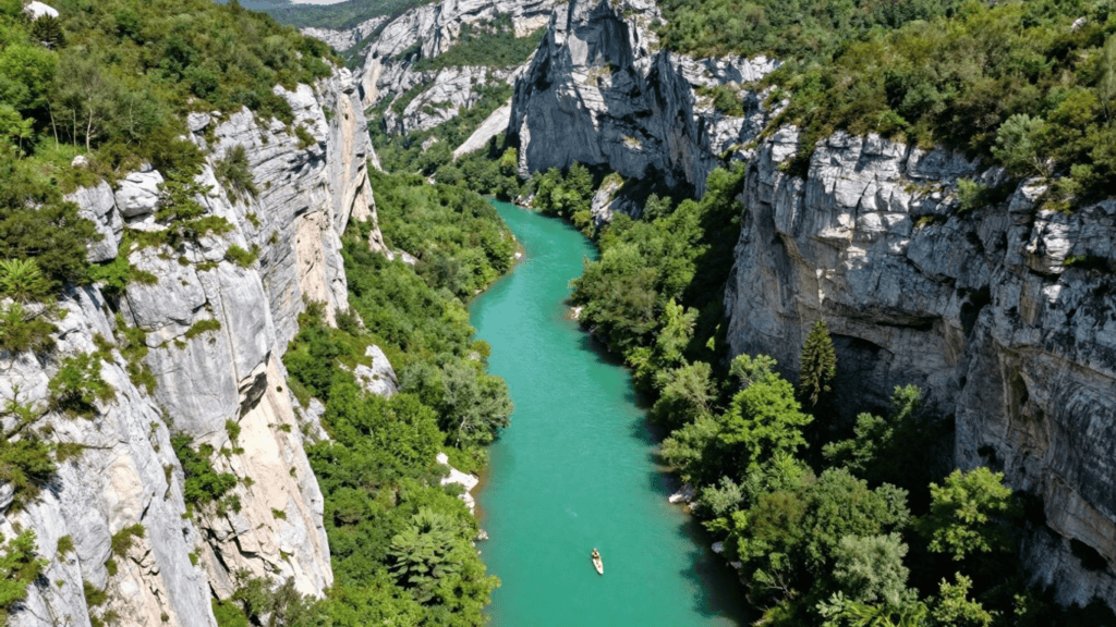 Aerial view of a turquoise river winding through steep limestone cliffs with dense green trees on both banks and a small boat paddling in the center.
