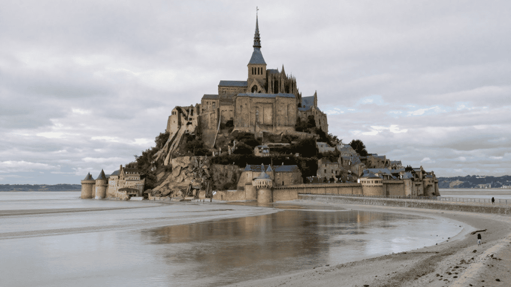 Mont Saint-Michel: a stone island abbey with a tall spire atop a rocky hill, connected by a causeway under an overcast sky.