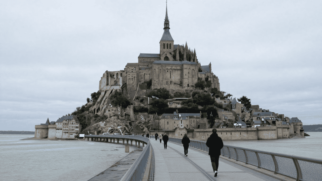 Medieval abbey on a rocky island connected by a long causeway, with a walkway and tourists approaching from the foreground on a cloudy day