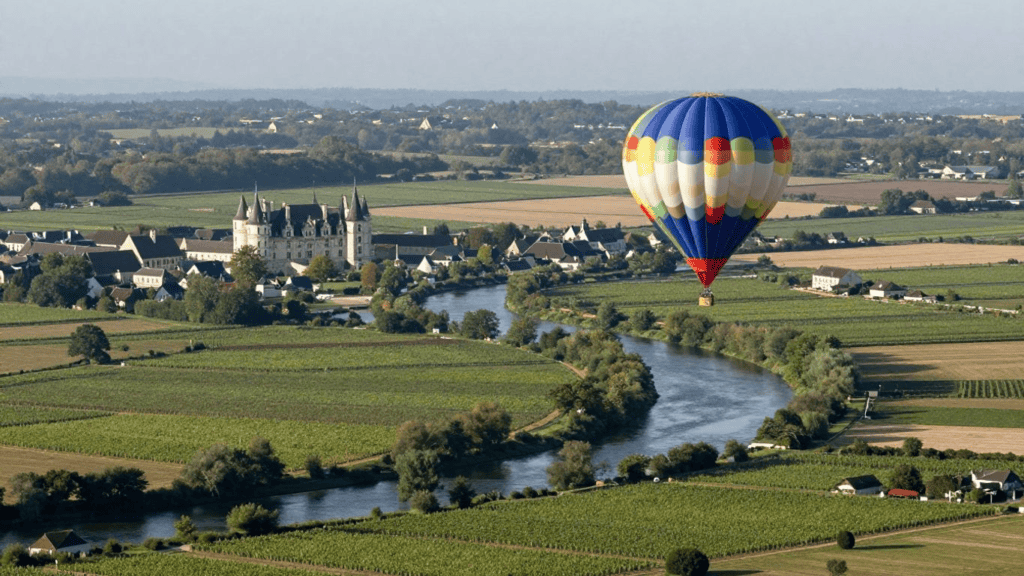 Colorful hot air balloon floating over a river valley, with a castle-like building and green farmland nearby.