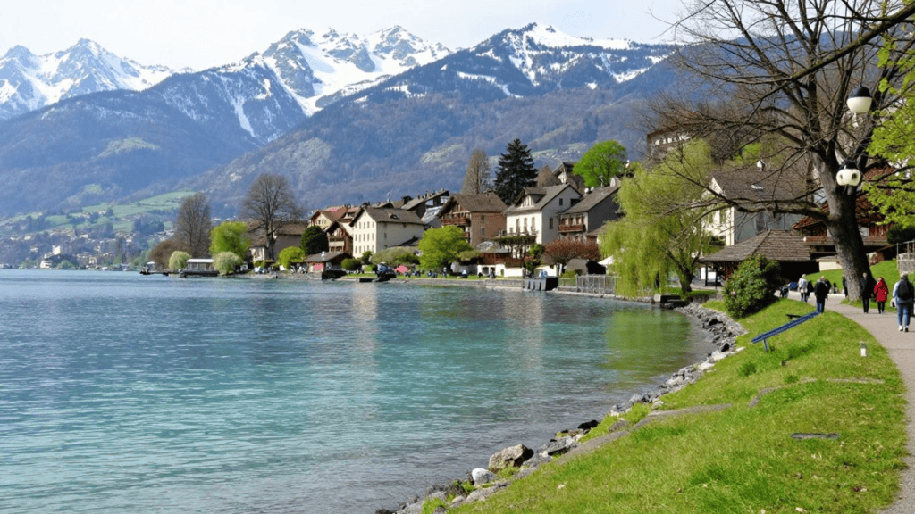 Lakeside town with pastel houses along a clear blue lake and snow-covered mountains in the distance.