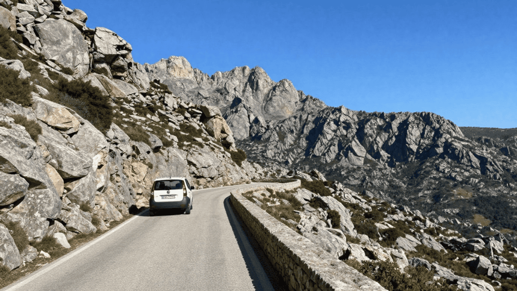 White SUV travels a narrow mountain road flanked by rocky cliffs and a stone barrier on the right under a clear blue sky.