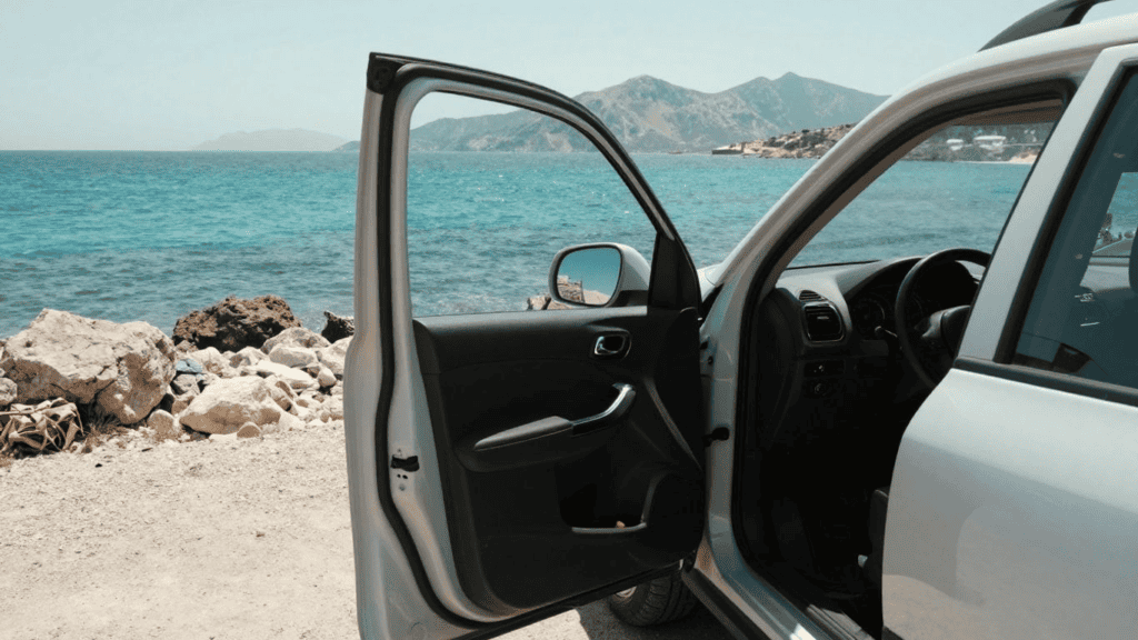 Open car door by a rocky beach with turquoise sea and distant mountains in the background.