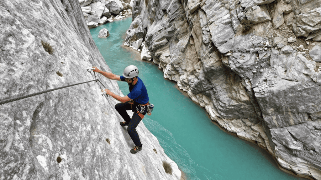 Man wearing helmet climbing a steep gray rock face above a turquoise river using a rope and harness.