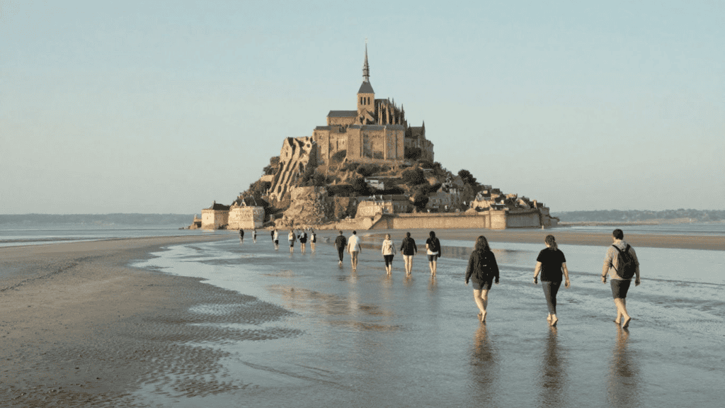 Tourists walking on a wet beach toward Mont Saint-Michel island under a clear sky.