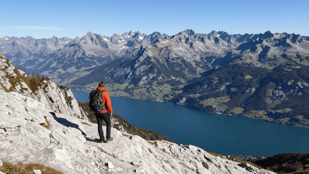 Hiker in an orange jacket with a backpack stands on a rocky slope, overlooking a blue lake and snow-capped mountains.